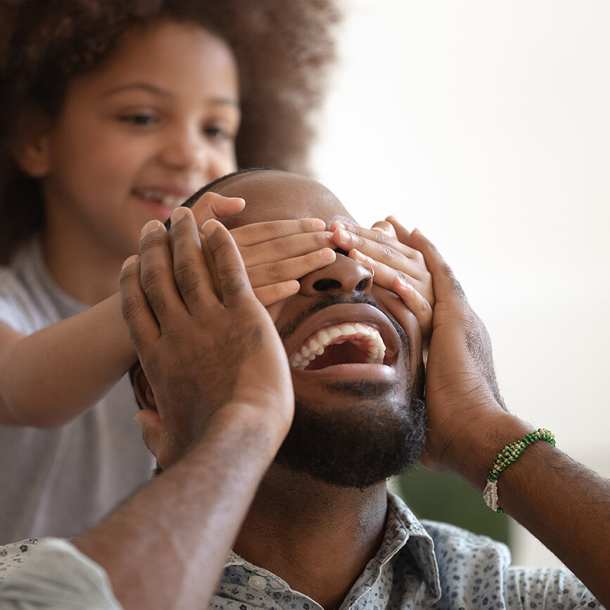 Cute mixed race ethnicity preschooler girl close dad eyes playing in funny game spend weekend at home together, smiling african American father and little daughter have fun engaged in family activity